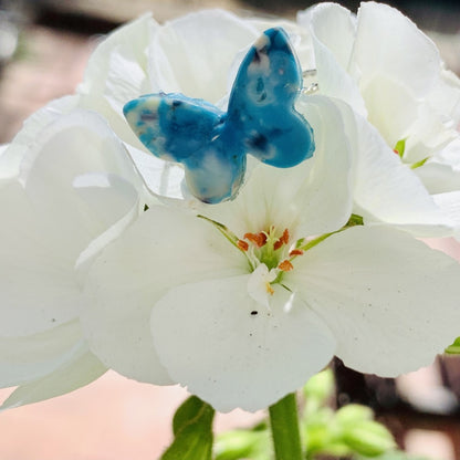 Resting on a bright white cluster of geranium blooms is a ring made from recycled 3D prints. It is in the shape of a butterfly with teal, blue, white, and black colors intermixed to be speckled like granite.