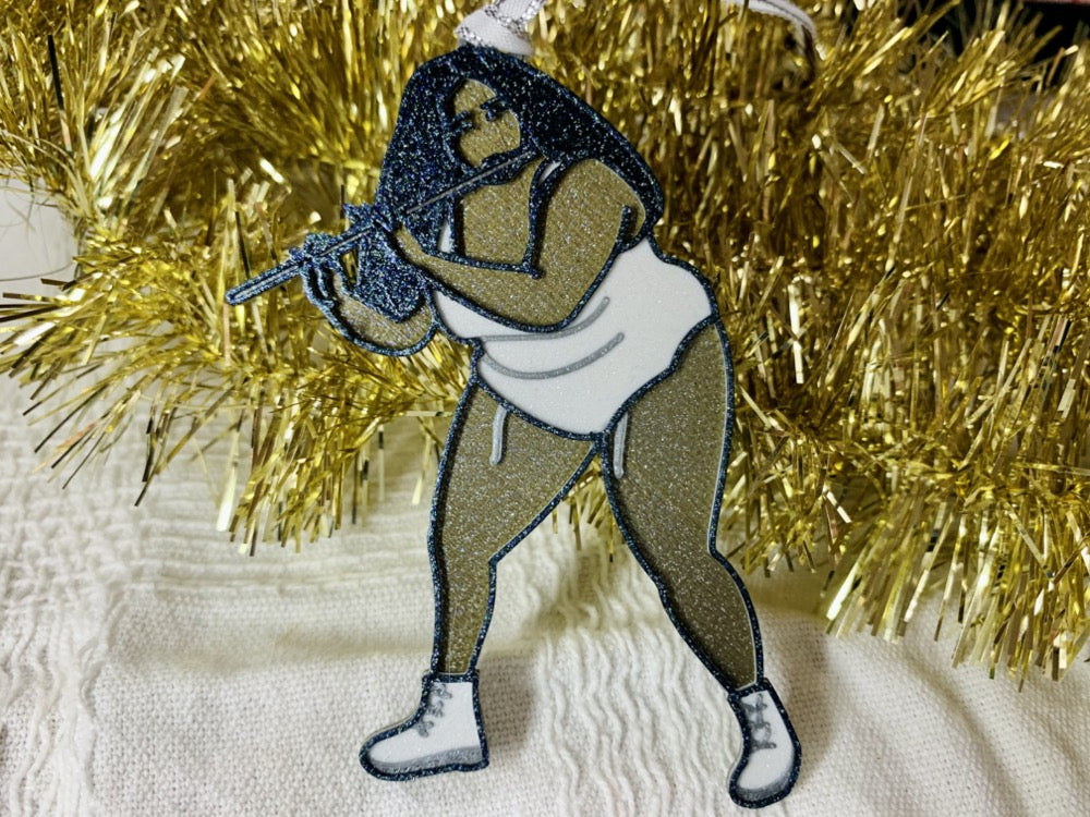 On a white fabric background there is an R+D 3D printed ornament resting on a pile of gold garland. The ornament is shaped like Lizzo while she plays her flute. She is gold, silver, white, and black and the entire ornament has been covered in glitter to shimmer in the light.