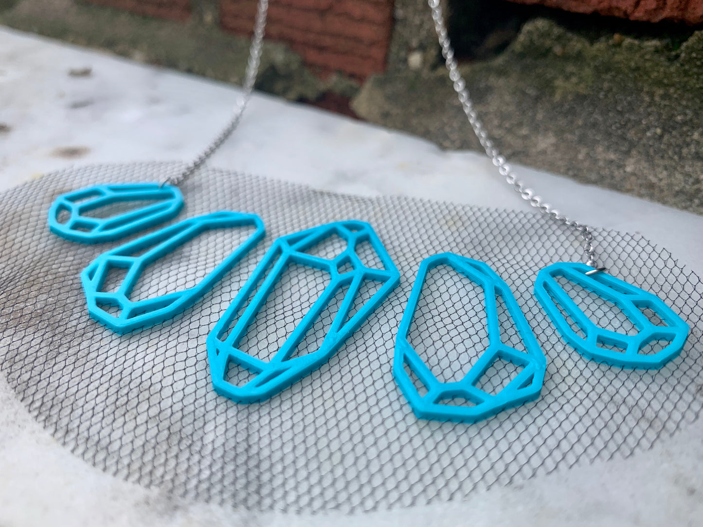 On a white marble background there is a R+D necklace. It is teal eco friendly filament in the shape of five crystals. They don't touch but are embedded with a black tulle fabric to stay together and look like they are floating when being worn.