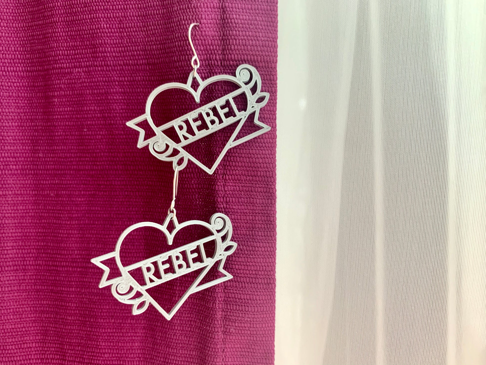 There are two silver earrings hanging from a purple curtain. The earrings are 3D printed and shaped like a heart tattoo. There is a heart, with a banner twisting around it, and two roses at the corners. The banner reads rebel and can be personalized. 