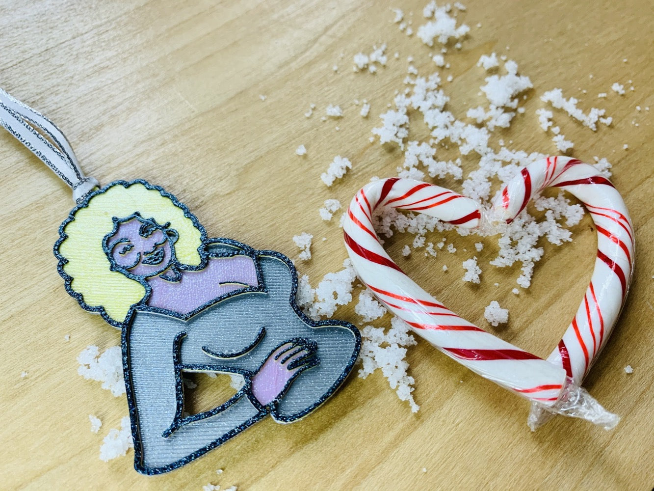 On a wood background with white snow and red and white candy canes is a R+D 3D printed ornament. This ornament is shaped like Dolly Parton. You can see one hand resting on her hip as she wears a silver top. Her trademark neckline and big blond hair is also visible with a smile. The entire ornament is covered in glitter to be able to shimmer and shine in the light.