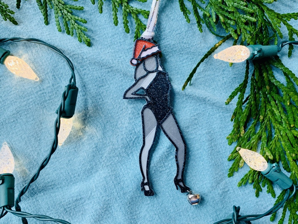 On a bright blue background, surrounded by greenery and a string of Christmas lights, is a R+D 3D printed ornament. It is shaped like beyonce from her Single ladies video, but also has her wearing a red and white santa hat. She is otherwise black and silver. 