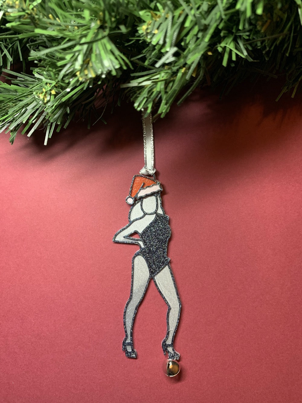 Hanging from a holiday wreath is a ornament that looks like Beyonce from her Single Ladies video: She is posing with her hand on her hip and one leg jutting out to the side. She is wearing a red and white santa hat and a black lone shoulder leotard. She is wearing black strappy heels and a small jingle bell hangs from one of her feet. The entire ornament is covered in a shimmery glitter that will catch all the lights of your holiday celebration. 