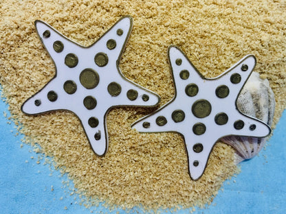 In the foreground are two R+D earrings. They are shaped like star fish and are white with gold outlines. There are gold circles that stretch out on each leg of the starfish. They are laying on sand and shells and a bright blue background is visible.