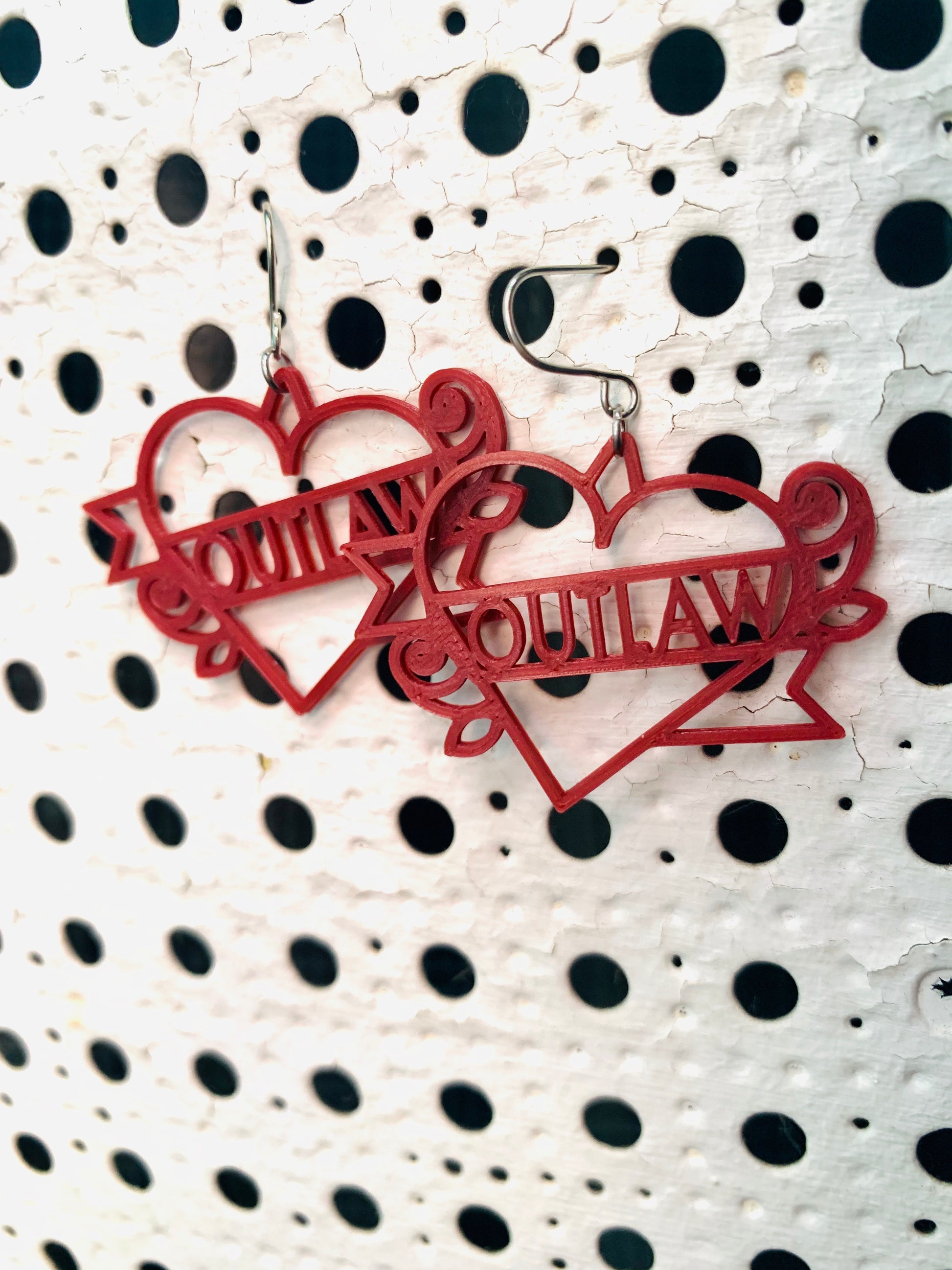 Two earrings are hanging off of a background with white paint and holes across it. The earrings are a dark merlot red and shaped like heart tattoos--they are  hearts with a banner twisting around it and two roses. The banner reads outlaw, but can be personalized to any word. They are 3D printed in an eco friendly filament. 