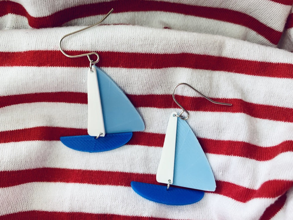 On wrinkled red and white striped fabric, there are two earrings shaped like sailboats. There is a thin white sail, a light blue sail, and a cobalt blue hull. 