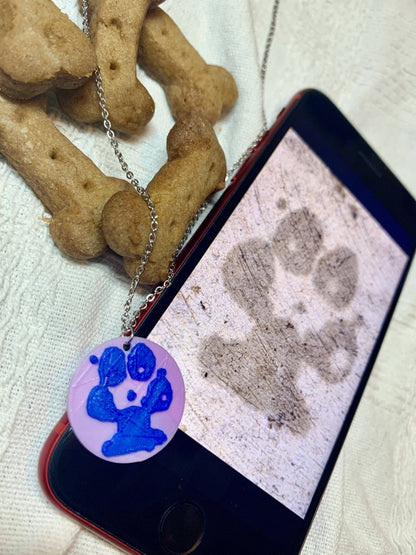 Shown laying on a white fabric is a phone, a necklace with a 3D pritned pendant, and two dog bone shaped treats. The pendant is a 1 inch light purple circle with a blue paw print on it. The picture on the phone is of the same paw print and was used to create the custom print.
