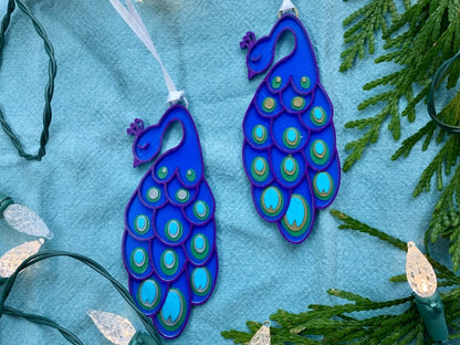 On a bright blue fabric background and surrounded by evergreen sprigs and Christmas lights are two R+D 3D printed peacock ornament. It has a blue body, with purple outlines. On the long elegant feathers are green, gold, silver, and teal accents.