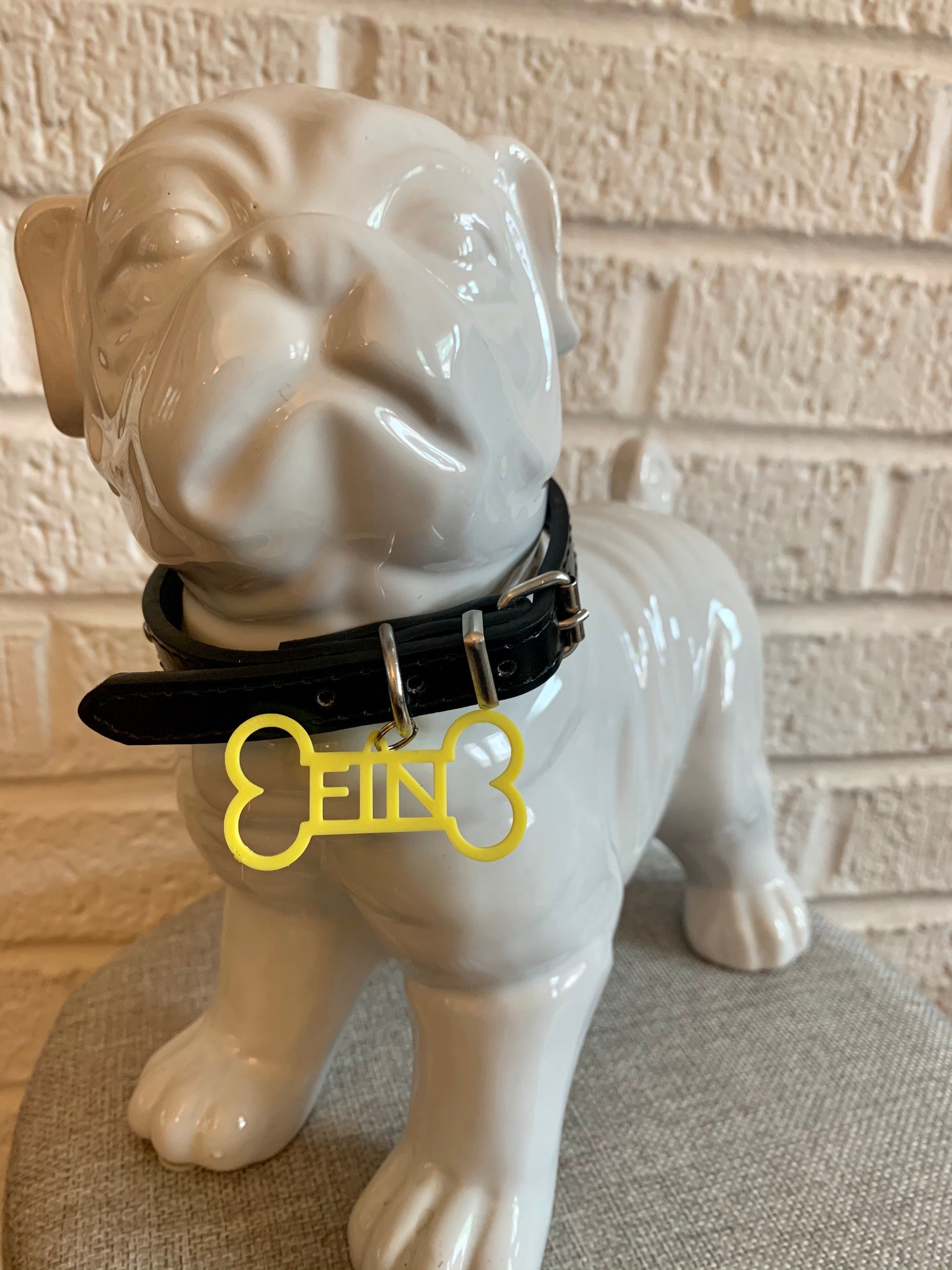 In front of a white brick wall and on a grey cushion is a ceramic bulldog statue. It is wearing a black collar and has a yellow dog tag. The dog tag is 3D printed in the shape of a dog bone with the name FIN in the middle. The name can be personalized to any name you would like.