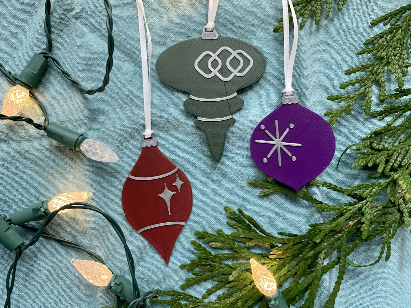 On a bright blue background with evergreen branches and a string of lights are a set of 3 R+D 3D printed ornaments. They are all designed to look like vintage baubles that would have commonly been found on Christmas trees in the past. They each have silver accents of stars and shapes. One is merlot red, one is dark green, and one is purple.
