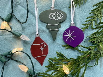 On a bright blue background with evergreen branches and a string of lights are a set of 3 R+D 3D printed ornaments. They are all designed to look like vintage baubles that would have commonly been found on Christmas trees in the past. They each have silver accents of stars and shapes. One is merlot red, one is dark green, and one is purple.