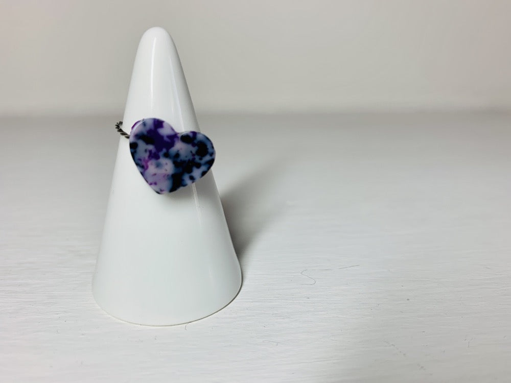 On a white background is a ring holder with a single cast ring resting on it. The ring is a smooth heart shape with a twisted metal band. The ring is cast from recycled 3D prints in shades of purple, black and white to create a marbled or granite like appearance.