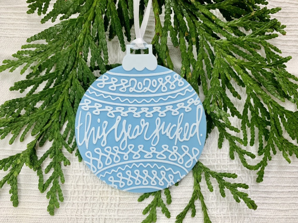 On a white fabric background there are sprigs of everygreen and a R+D 3D printed ornament. The ornament is shaped like a traditional bulb. It is light blue with white drawings and script on it. Worked into the doodles are the year 2020 and then the words, "this year sucked".