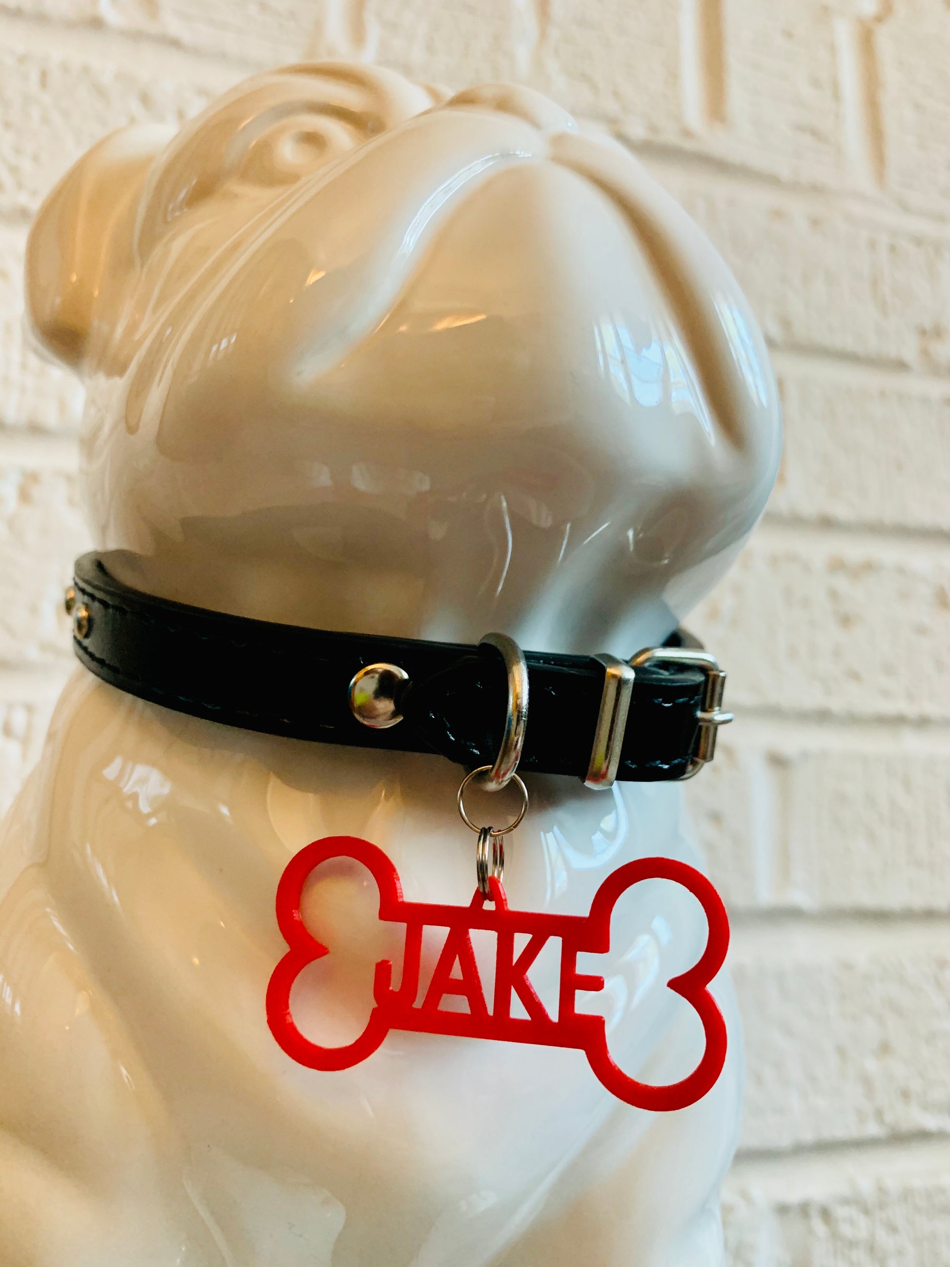 In front of a white brick wall and on a grey cushion is a ceramic bulldog statue. It is wearing a black collar and has a red dog tag. The dog tag is 3D printed in the shape of a dog bone with the name JAKE in the middle. The name can be personalized to any name you would like.