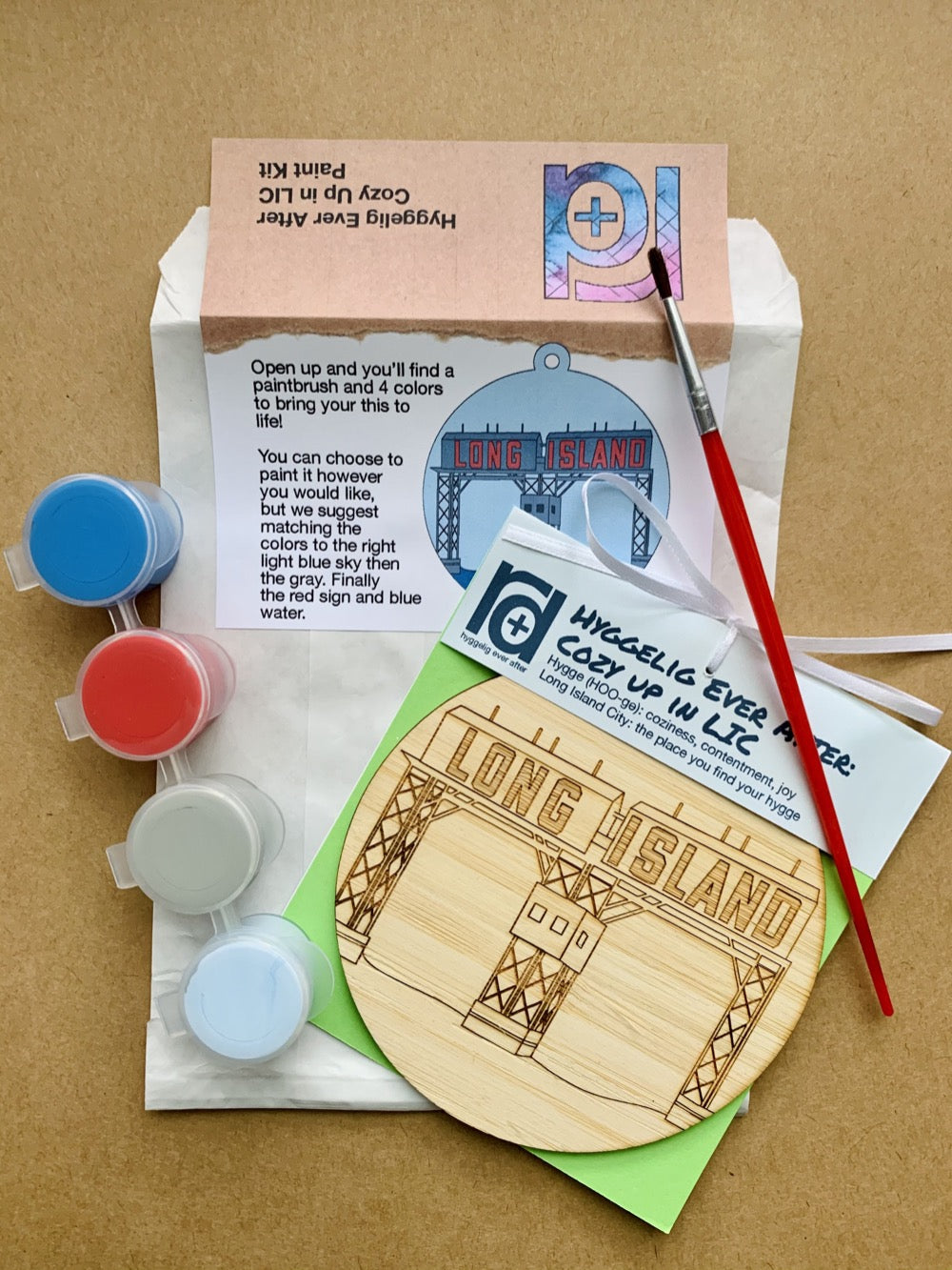 Shown laying on craft paper is the packaging and contents of this DIY Paint Kit. There is a paper envelope with instructions, a paintbrush, a set of 4 paint wells and a wall hanging with a laser cut NYC landmark.