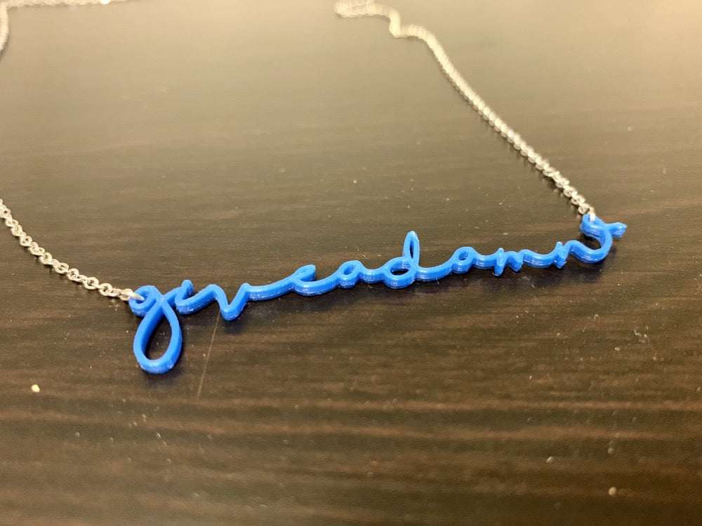 On a dark wood surface is a 3D printed pendant. In a modern cursive script it says give a damn  in a classic cobalt blue color.