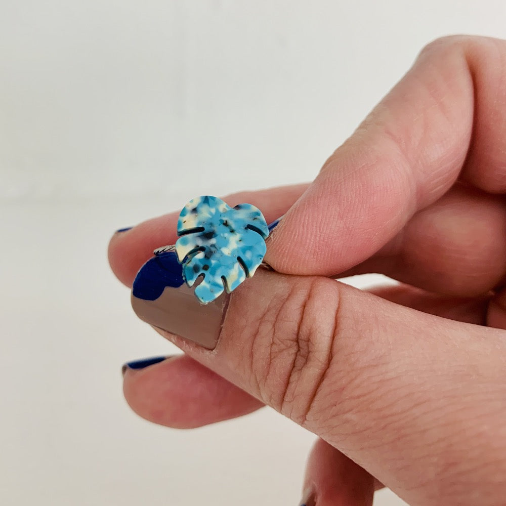 A hand is holding a ring on a white background. The ring has a thin twisted metal band and a charm at the front. The charm is the shape of a monstera leaf and is made from recycled 3D prints in teals, blues, white, and black colors to give it the look of granite or speckled marble.