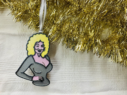 On a white fabric background with gold tinsel garland is a R+D 3D printed ornament. This ornament is shaped like Dolly Parton. You can see one hand resting on her hip as she wears a silver top. Her trademark neckline and big blond hair is also visible with a smile. The entire ornament is covered in glitter to be able to shimmer and shine in the light. 