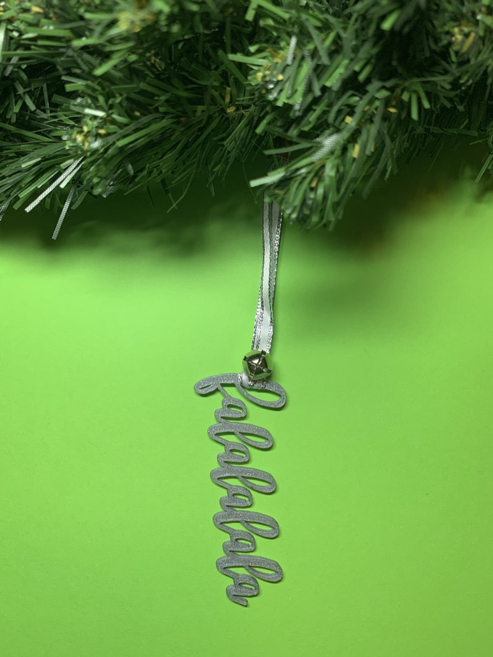 On a bright green background and hanging from a green wreath is a 3D printed R+D ornament. It is a cursive text with a jingle bell and covered in glitter to make it shimmer and shine in the light. This ornament reads, falalalala.