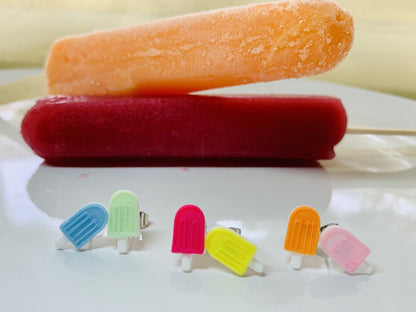 In the foreground are three sets of R+D stud earrings. They are mismatched, but complementary colors: One set is light blue and mint green. The next is hot pink and yellow. The third is orange and light pink. In the background are two actual popsicles. One a deep raspberry red and the other a tangerine orange.