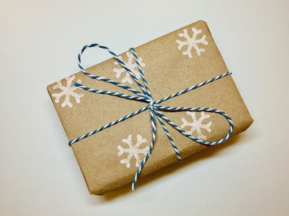 In the center of the photo is a wrapped gift. The wrapping paper is 100% recycled craft paper with white snowflakes stamped on. There is blue and white striped twine tied around the box and in a bow.