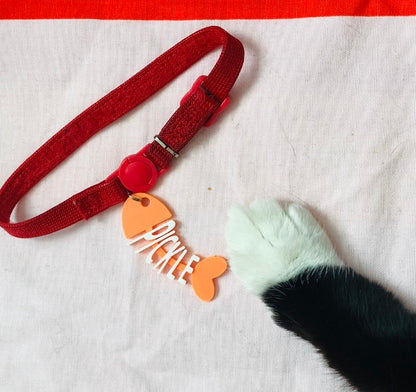 A black and white cat's paw is reaching into the frame. There is a red collar with a tag on it. There is a cat tag that is shaped like a cartoon fish that has been eaten. The head, tail, and backbone is bright orange. Where the bones would be, there are letters to form the name Pickle in white.