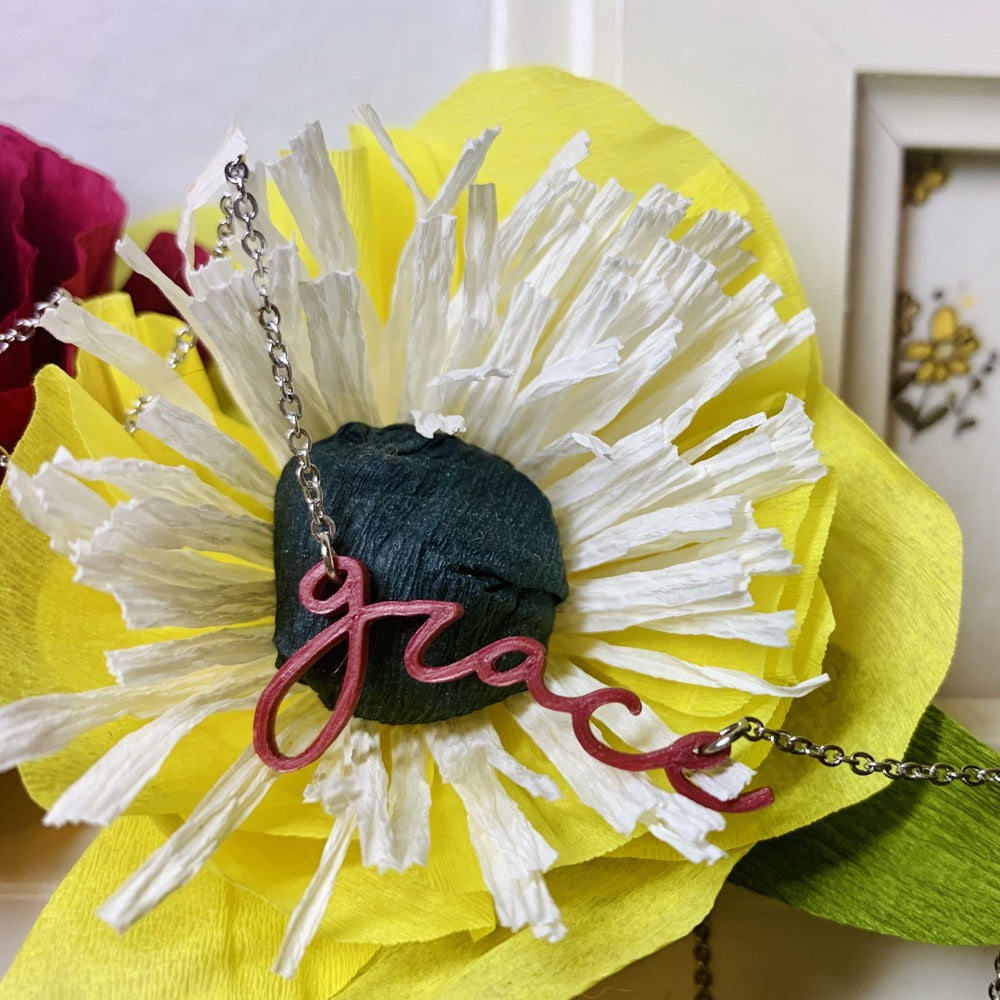 Hanging on a paper flower that is yellow, white, and black is a 3D printed pendant. In a modern cursive script it says grace in a dark red color.