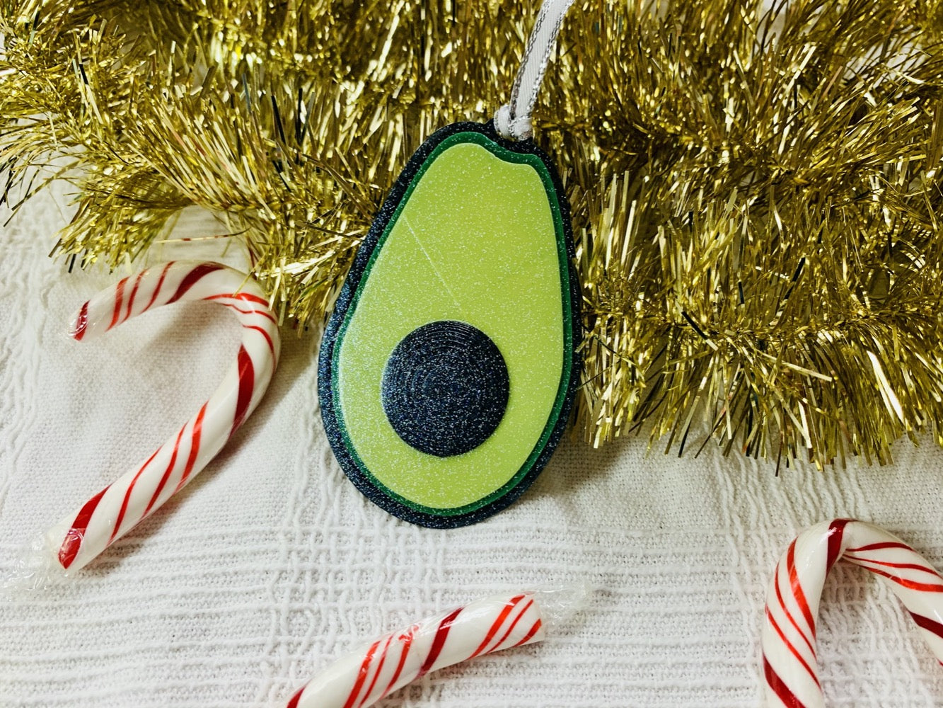 On a white fabric and gold garland background is a R+D 3D Printed ornament. It is shaped like an avocado that has been split open with a black peel, bright green highlights and lime green in the center. There is also a raised black pit in the middle. The entire ornament is covered in glitter so it will shine and shimmer in the light. To the side there are also red and white candy canes.