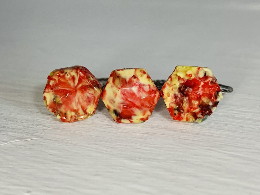  There are three rings side by side on a white background. They each have a hexagon shape with a twisted metal band. They are made from recycled remnants of plant based filament which gives them a speckled look like granite. The first ring has a smooth surface on the top, the middle a faceted texture and the third a rough texture like a geode being formed. These rings are all using red, yellow, orange, and black filaments to make these one of a kind pieces.