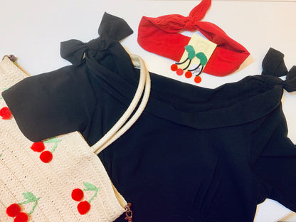 An outfit is laid out -- In the center is a boat necked dress with bows at the shoulders. There is a bright red kerchief, R+D earrings shaped like cherries, and a straw bag over the shoulder with cherry pom poms sewn on.