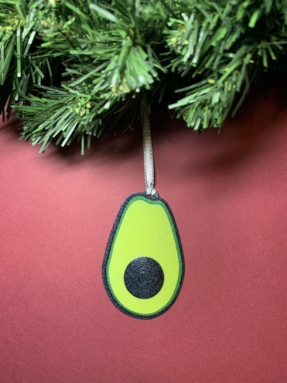 On a red background and hanging from a green wreath is a R+D 3D Printed ornament. It is shaped like an avocado that has been split open with a black peel, bright green highlights and lime green in the center. There is also a raised black pit in the middle. The entire ornament is covered in glitter so it will shine and shimmer in the light.