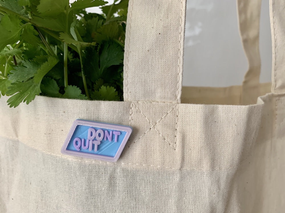 On a cream reusable tote bag, there is a 3D printed pin. It is a hexagon shape in light blue plant based filament with the words DONT QUIT in light purple. There is a hidden message that is opaque away from UV, but will turn hot pink in sunlight and read DO IT. 