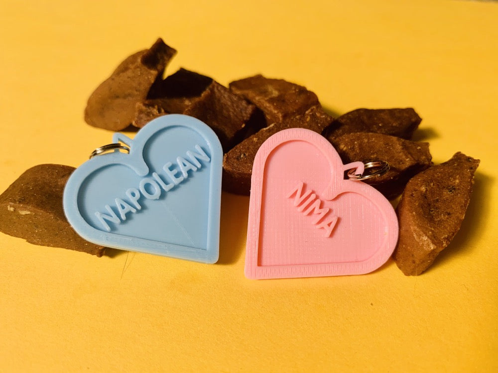 On a bright yellow background is a pile of fish shaped cat treats. In the foreground are two 3D printed pet tags. They are in the shape of a heart with the pets names in the center. One is light blue with the name NAPOLEAN the other is light pink with NIMA in the middle. They are able to be personalized with different colors and names. 