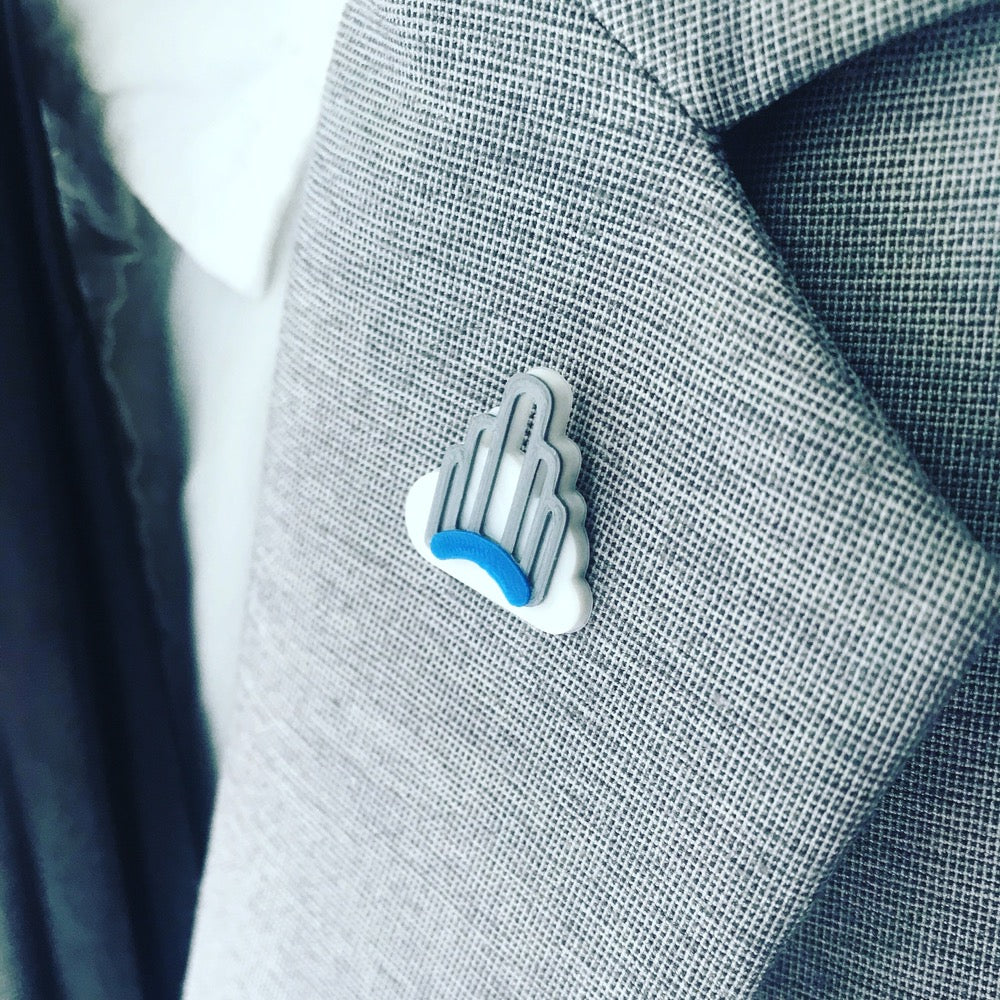 The magnetic 3D printed pin is show on the lapel of a grey suit worn with a white shirt and black tie. The pin is a whit cloud in the background of a silver and blue art deco arch that stands in the neighborhood of Sunnyside, Queens, NYC. 