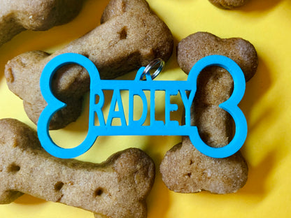 Pictured on a yellow background with dog treats shaped like bones is a 3D printed dog tag. The tag is bright teal and also shaped like a dog bone and have the name RADLEY in the middle. It can be customized to any pets name.