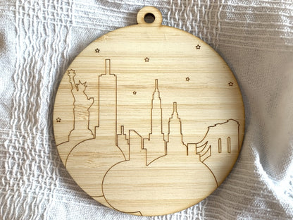 Shown on a white fabric background is a laser cut wall hanging. It is cut from bamboo, a fast growing and sustainable grass. Etched in it a picture of the New York City Skyline with the Statue of Liberty, Empire State Building, Chrysler Building and Brooklyn Bridge all visible. Each piece can be painted like a paint by number or left plain. It can hang from your Christmas Tree or on you wall all year round.