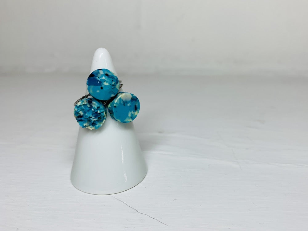 On a white background and slipped on a white cone ring holder are three rings cast from recycled 3D prints. The rings are each in a circular shape. One has a smooth surface, another a faceted surface, and finally one like a geode forming. They each have teal, blue, white, and black colors intermixed to be speckled like granite.