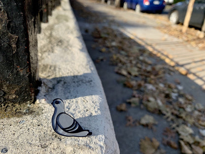 Resting on the sidewalk outside a park is a close up of a 3D printed pin. It is silver and black, shaped like a pigeon and 3D printed with plant based materials.