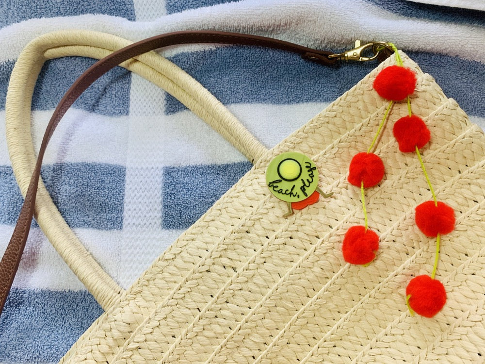 Pinned to a light straw bag is a R+D pin. It shows someone in a red bathing suit resting back on their hands. They are wearing a large brimmed yellow hat that has beach please written on it in a black script. Hanging off the bag are bright red pom poms. This is all  resting on a blue and white striped towel. 