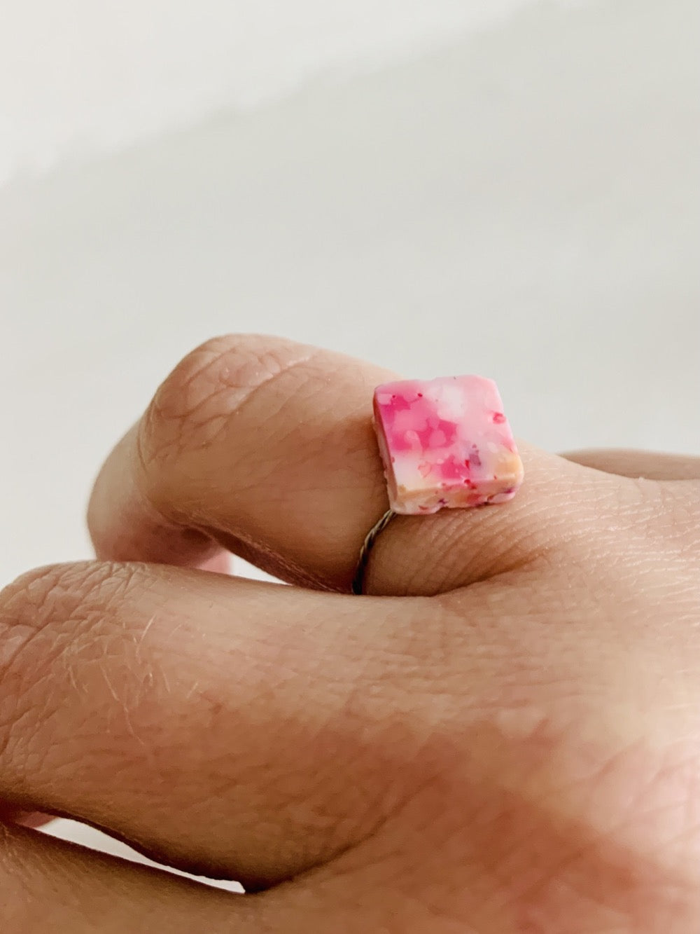 Shown on a person's ring finger and in front of a white background, there is a ring with a cast charm on a thin twisted band. The charm is cast from recycled 3D prints in shades of pinks, yellow, orange, and white. It has the look of granite or marble. 