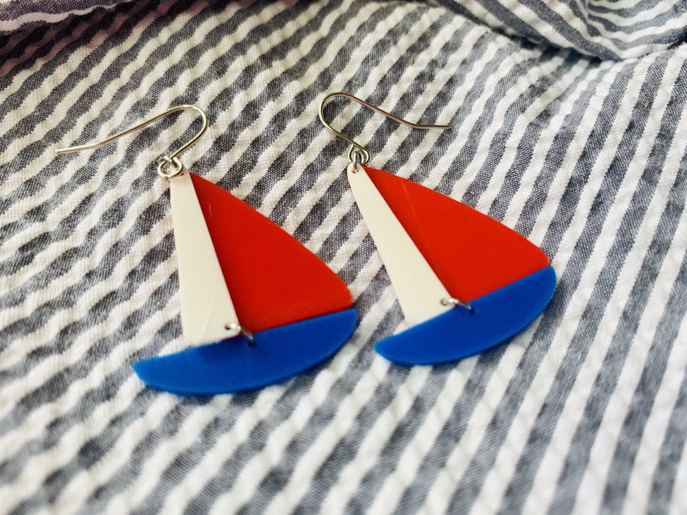 Two earrings are laying on seersucker fabric. The earrings are shaped like sailboats with a thin white sail, a bright red sail and a cobalt blue hull. 
