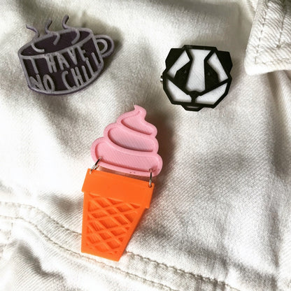 Three plant-based 3D printed pins on a white denim jacked. One is shaped like a ice cream cone with a solid pink ice cream swirl above an orange sugar cone. Above these are a black and white geometric panda pin. There is also a tea cup shaped pin that shows as purple and a opaque white color. It has the words "I have no chill" written on it in the opaque white. In direct sunlight, this will turn bright hot pink. 