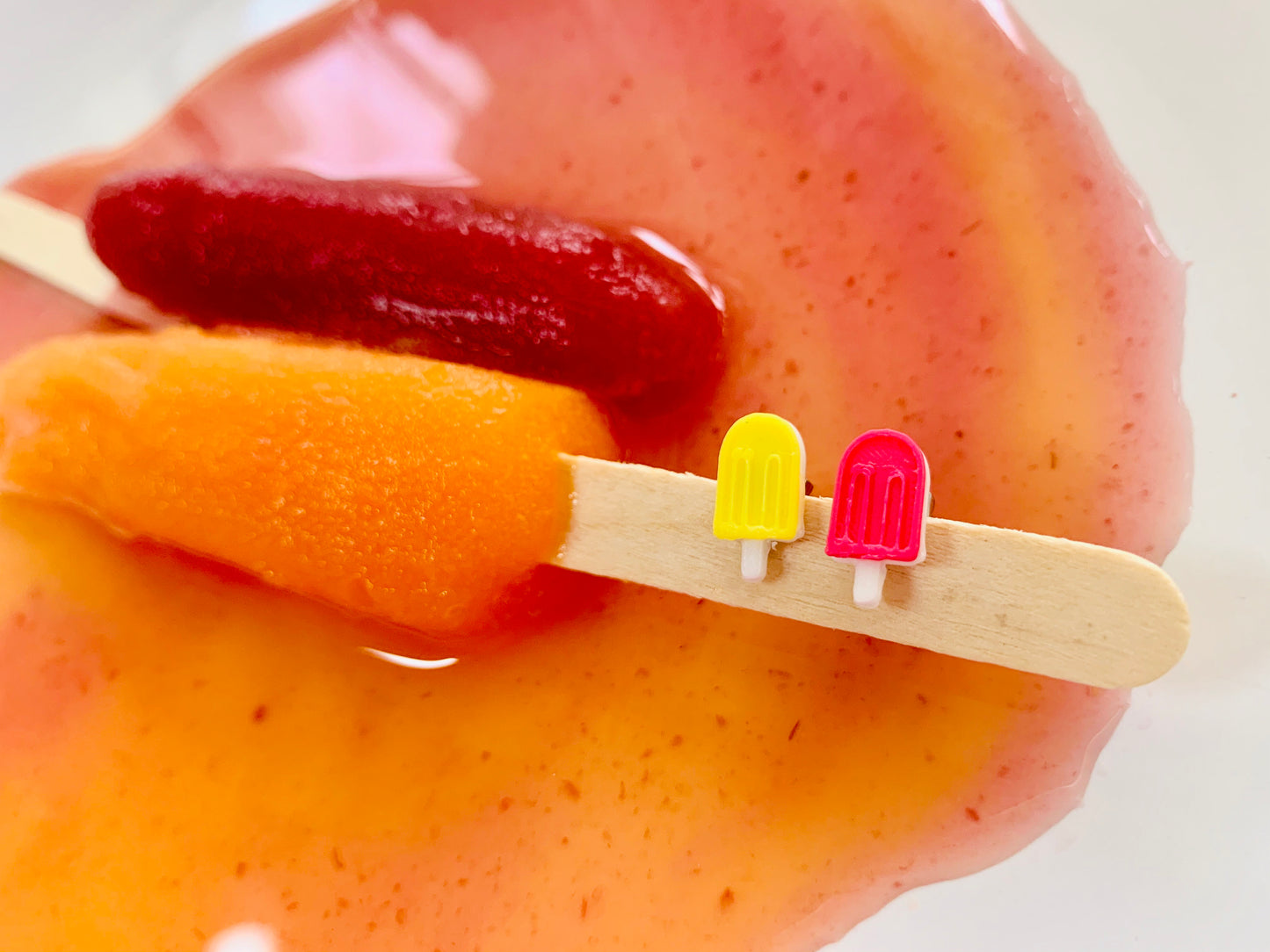 There are two mostly melted popsicles in a pool of juice. One is a rich dark red, the other is a bright  orange. On the wooden popsicle stick are two small  3D printed earrings from R+D. They are mismatching, but complementary colors. One is yellow, the other is bright pink. Both have white sticks. 