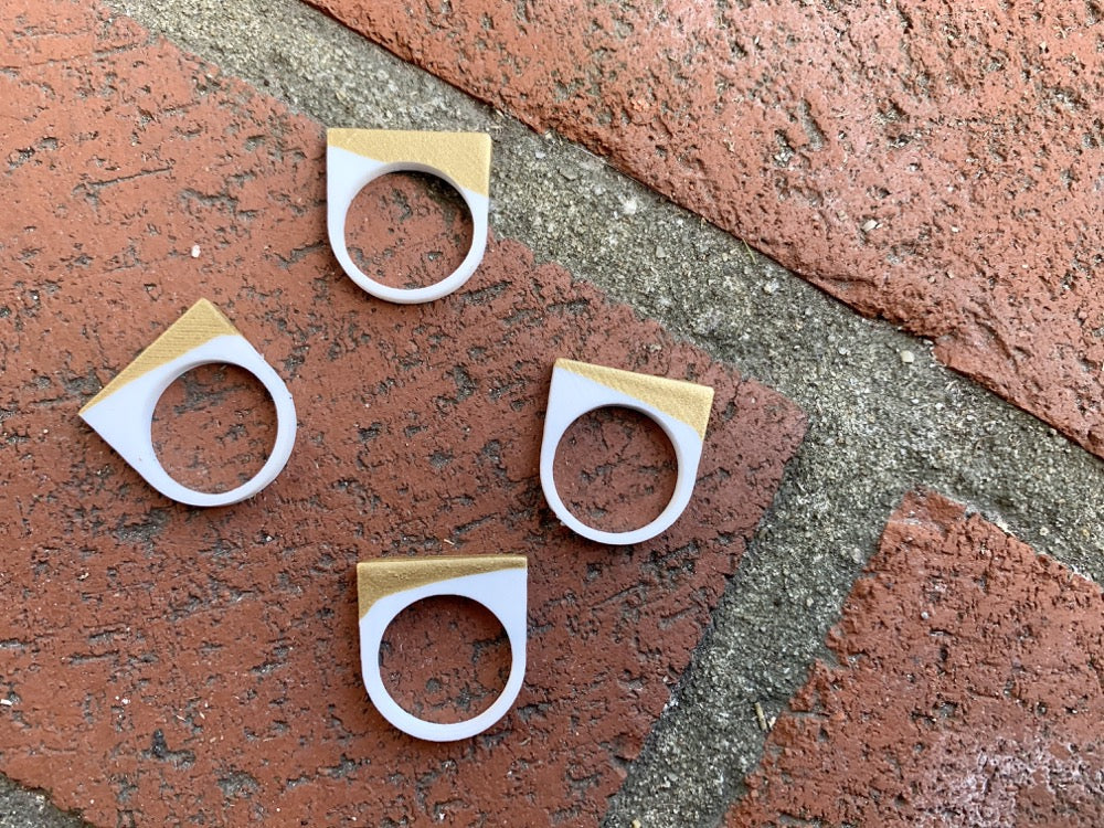 Laying across a brick pathway are four 3D printed rings. They are each smooth circle bands that extend into a square shape above the finger where worn. They are printed in a white plant based filament and dipped into a gold paint to give a unique addition across the top of each ring. 