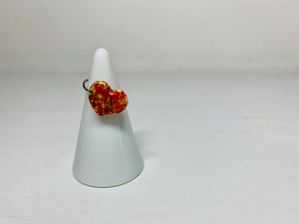 On a white background and slipped on a white cone ring holder is a ring that is cast from recycled 3D prints. The ring is in a heart shape with red, yellow, orange, and black colors intermixed to be speckled like granite.