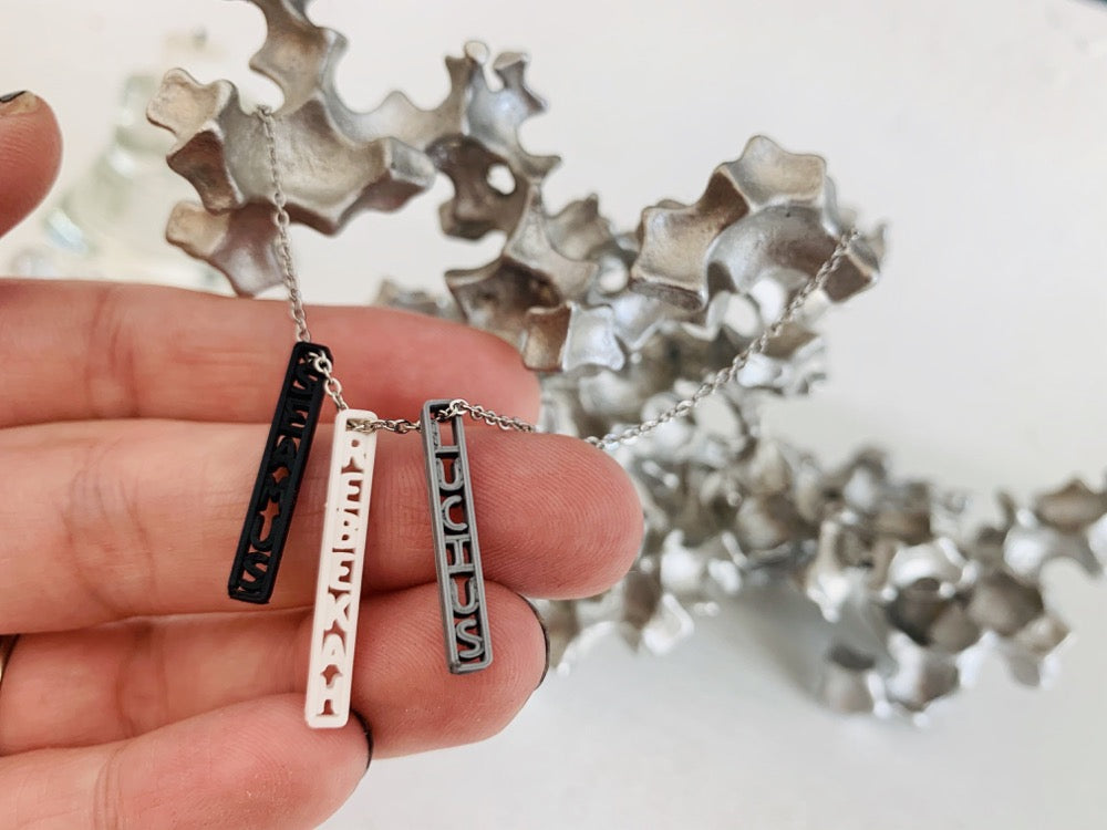 Shown on a white background with an aluminum sculpture, there is a necklace with 3 3D printed pendants. There is a hand reaching in to show the sides of each pendant, in silver, white, and black. These are turned to the side to reveal three names: SEAMUS, LUCIUS, and REBEKAH. They can be customized to any name or word.