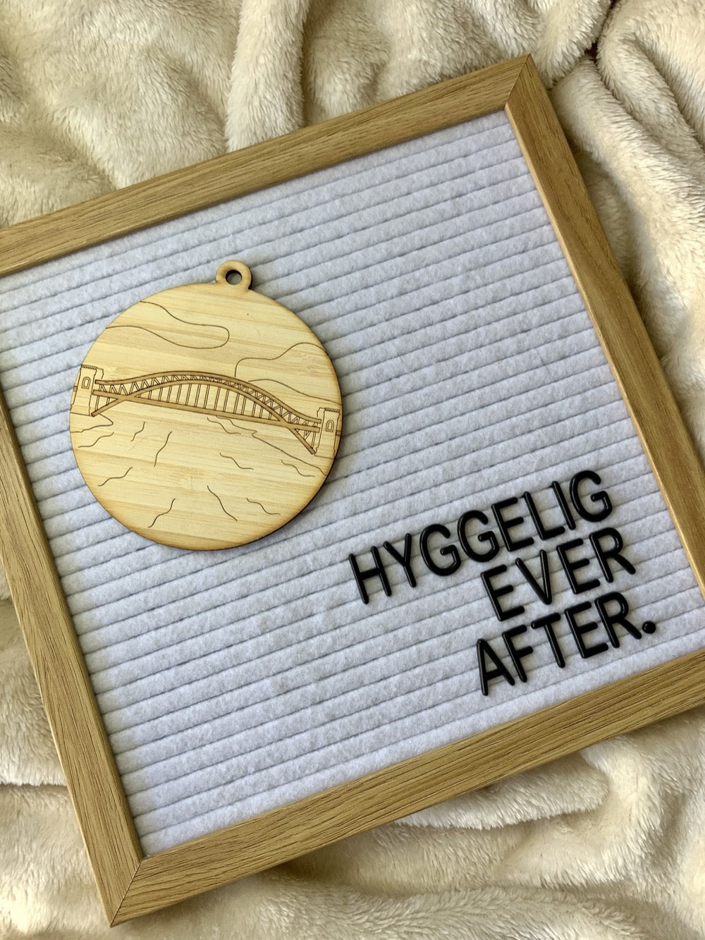 Shown laying on a plush blanket is a letter board that reads "Hyggelig ever after". Also on the letter board is a laser cut wall hanging. It is cut from bamboo, a fast growing and sustainable grass. Etched in it a picture of the Hell Gate bridge in Astoria. This bridge is an icon of the area and looked on from Asttoria Park. This can be used as a ornament in the holidays or a wall hanging all year round. It can be painted like a paint by number or left plain.