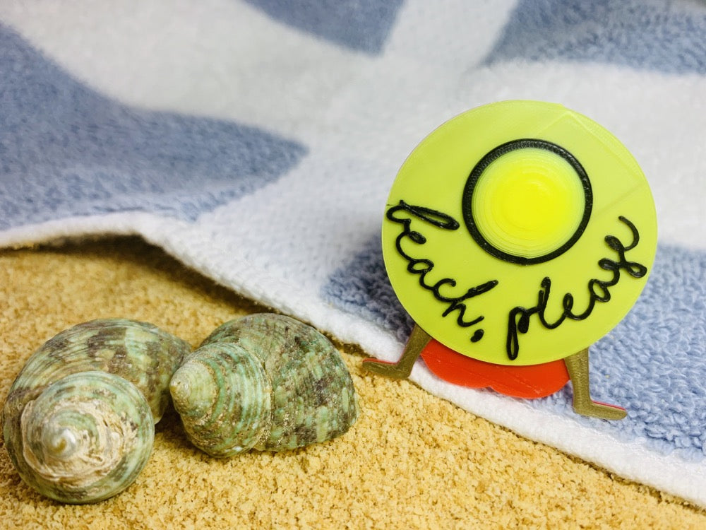 On the edge of a blue and white striped beach towel, next to sand and shells, is a 3D printed pin from R+D. The pin shows  someone wearing a red bathing suit and resting back on their hands. They are wearing a large, wide brimmed yellow hat that reads beach, please in a scripted black font. 