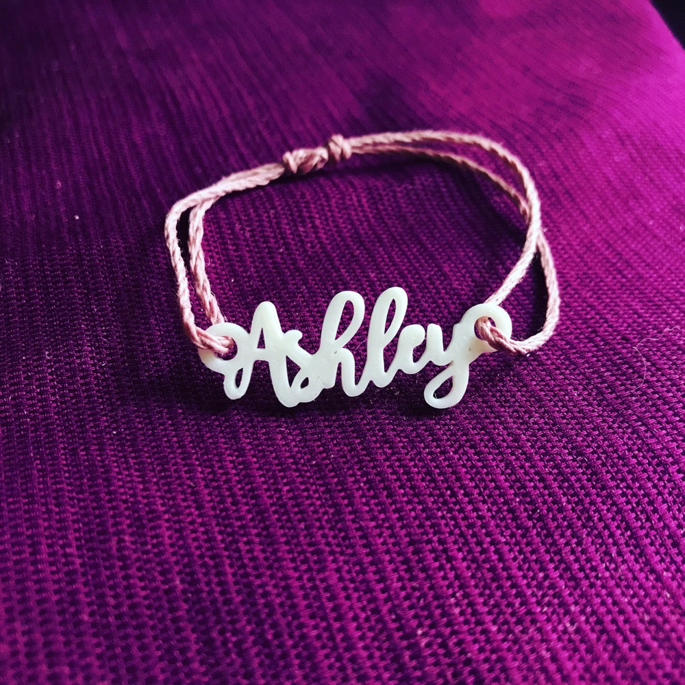 Shown on purple fabric is a bracelet with an adjustable cord. The charm in the center is 3D printed and reads Ashley in cursive script.