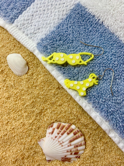 A light blue and white striped beach towel is covering sand. There are shells in the sand. On the towel are two R+D earrings. They are bright yellow and shaped like a bikini top and bottom -- the earrings are asymmetrical for each piece. On the bathing suit are white polka dots.
